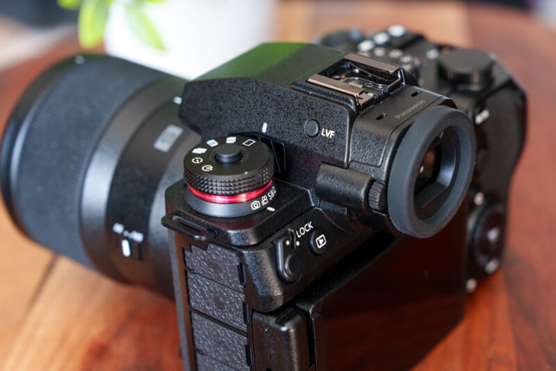 Close-up of a black DSLR camera placed on a wooden surface. The camera features a large lens, multiple control dials, a viewfinder, and a screen with a tilting mechanism. A blurred plant is visible in the background.