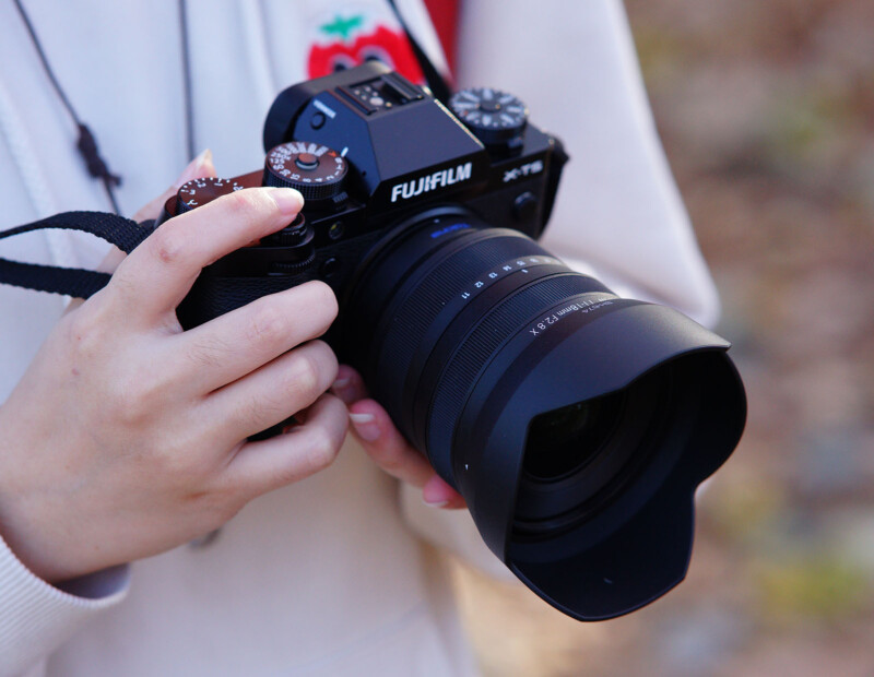 Person holding a Fujifilm X-T1 camera with a large lens, wearing a light-colored hoodie. Background is blurry, suggesting an outdoor setting.