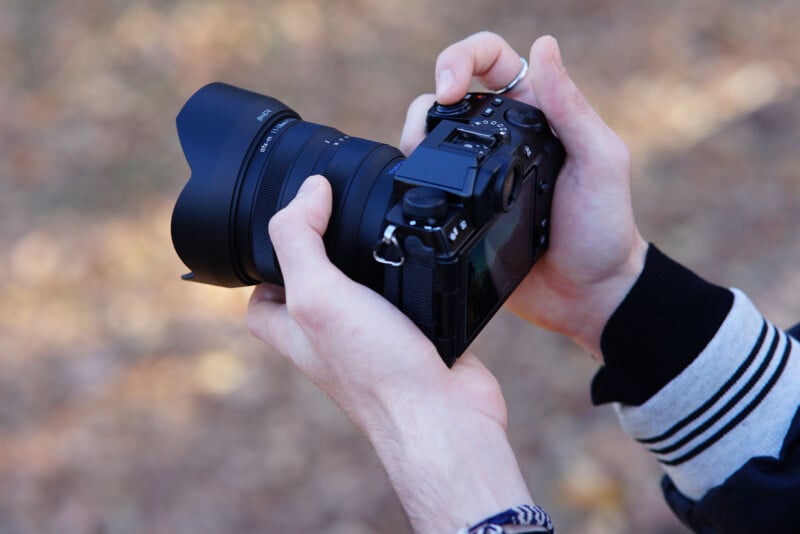 Hands holding a digital camera with a large lens, set against a blurred outdoor background. The person is wearing a black and gray jacket and a bracelet on the right wrist.