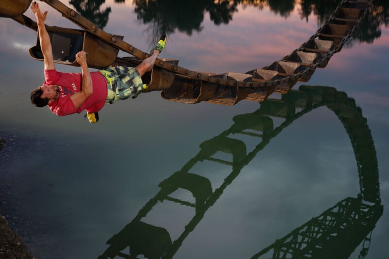 A person in a red shirt and checkered shorts hangs upside down from a roller coaster track above a body of water, with the track reflecting in the water. The sky is tinged with pink and blue hues.