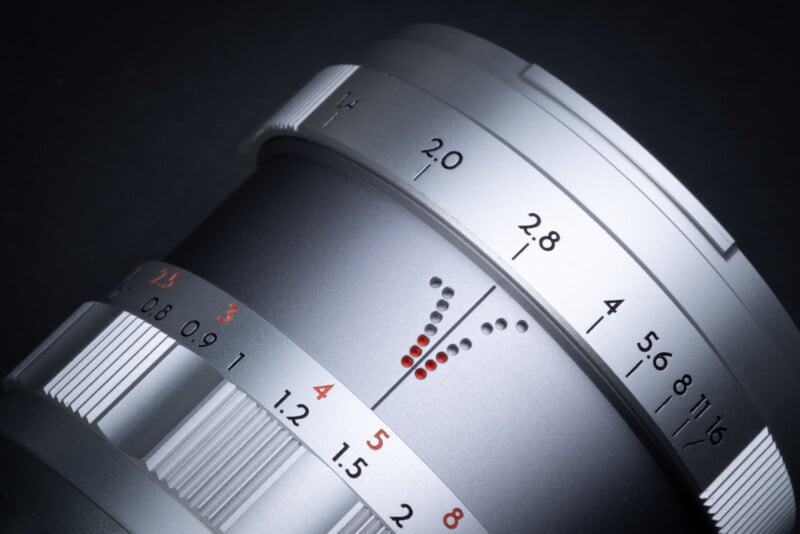 Close-up of a camera lens with focus and aperture markings. The lens features silver and black detailing, with numbers from 1.4 to 16 in black and red, indicating different aperture settings. The background is a dark gray.