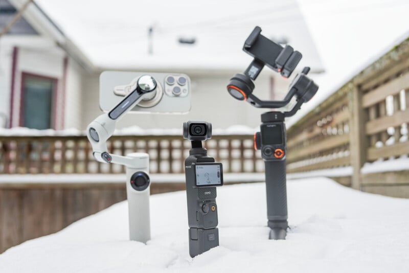 Three types of camera gimbals are placed upright in the snow: one holding a smartphone, one without any device, and a small handheld model in the foreground. A house and wooden fence are blurred in the background.