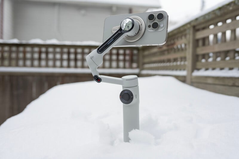 A smartphone mounted on a gray gimbal stands upright in the snow. The background features a wooden fence and a blurred building. The gimbal and phone appear stable despite the snowy setting.