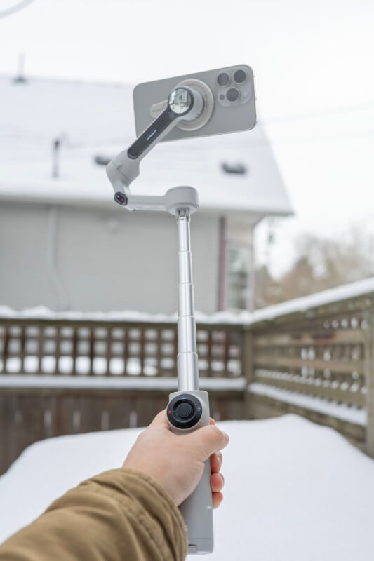 A person holds a silver selfie stick with a smartphone attached, aimed toward a snow-covered backyard. There's a wooden fence and a house in the background. The sky is overcast.