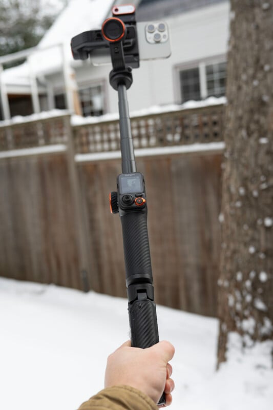 A hand holds a black selfie stick with a smartphone and an action camera attached. The background shows a snow-covered outdoor area with a wooden fence and a house.