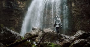 Person in a raincoat standing on moss-covered rocks, gazing at a large waterfall cascading down a rocky cliff. The surroundings are lush and misty, creating a serene and natural atmosphere.