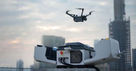 A drone hovers above a rooftop docking station in an urban environment. The background includes tall buildings under construction against a cloudy sky. The docking station appears open and contains electronic components.