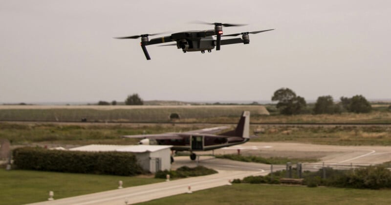 A drone is flying in the foreground with a small airplane parked on a runway below. The background features an open landscape with fields and overcast skies.