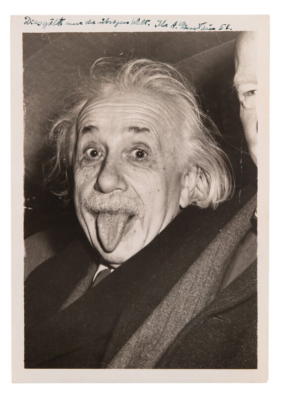 A black and white photo of an older man with wild white hair, a mustache, and a suit sticking his tongue out playfully. There is handwriting at the top of the photograph.