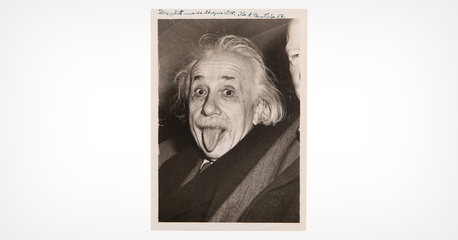 A black and white photo of an older man with wild hair sticking out his tongue. He is wearing a suit and a scarf. The background is plain, and part of a hand is visible in the corner. There is handwriting along the top edge of the photo.