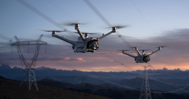 Two drones fly near power lines during a sunset. The sky displays shades of orange and violet, with mountain silhouettes in the background.