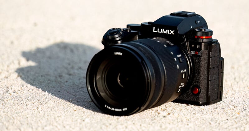 A Panasonic Lumix camera with a large lens sits on sandy ground. The camera is black with red accents and has "S1R" written on the grip area. The lens cap displays the Lumix logo, and the scene is lit by sunlight.