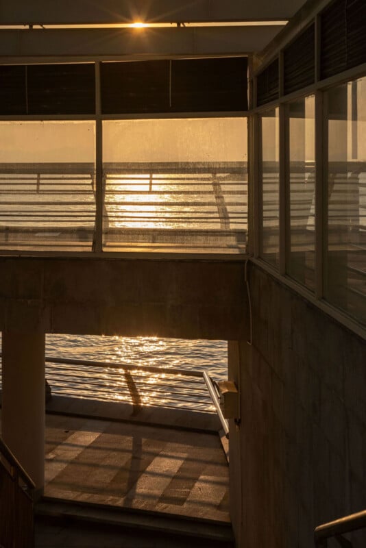 Sunlight filters through a window onto a stairway, casting shadows on the floor. The scene overlooks the ocean with shimmering reflections on the water, creating a warm and serene atmosphere.