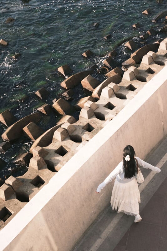 A person in a white dress walks along a concrete path beside a body of water with geometric, stone wave breakers. The person has long hair tied with a white accessory and holds a bag while extending their arms slightly.