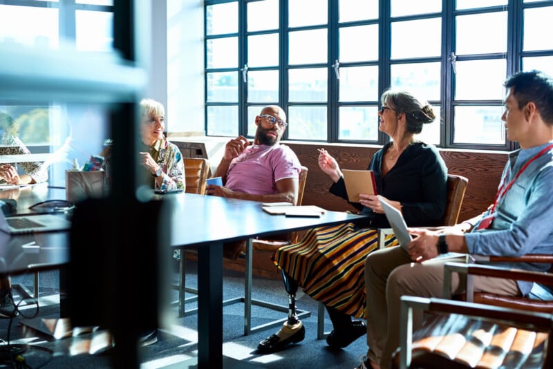 A diverse group of people sitting in a meeting room, engaged in discussion. One person has a prosthetic leg, and another holds a tablet. Sunlight streams through large windows in the background, creating a bright atmosphere.