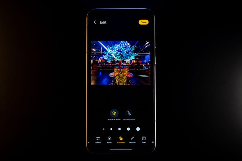 A smartphone displaying an image editing app. The screen shows a tree decorated with lights in a dimly lit room. Editing options are visible below the image, with buttons for adjusting, filtering, and text. The "Save" button is highlighted.