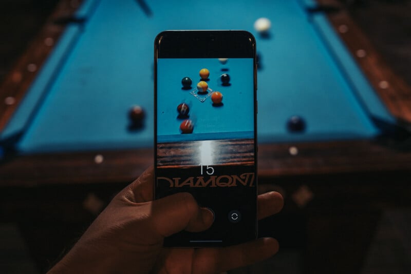 A hand holds a smartphone capturing a photo of a pool table. The screen shows the table with colorful billiard balls in focus. The actual pool table and balls are visible in the background, slightly blurred.