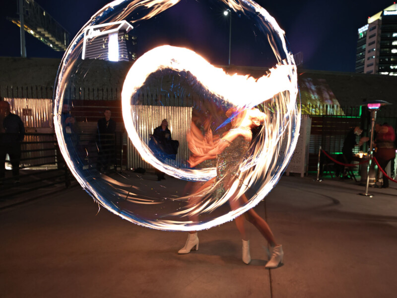 A performer spins a flaming hoop, creating a circle of fire in the air. The scene is set outdoors at night, with onlookers and tall buildings in the background. The performer wears a shiny outfit and white boots.