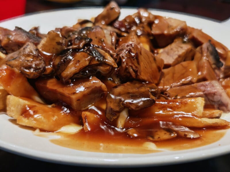 A plate of poutine topped with sliced roast beef and sautéed mushrooms, drizzled with rich brown gravy. The dish is served on a white plate, with crispy fries and cheese curds peeking through under the toppings.