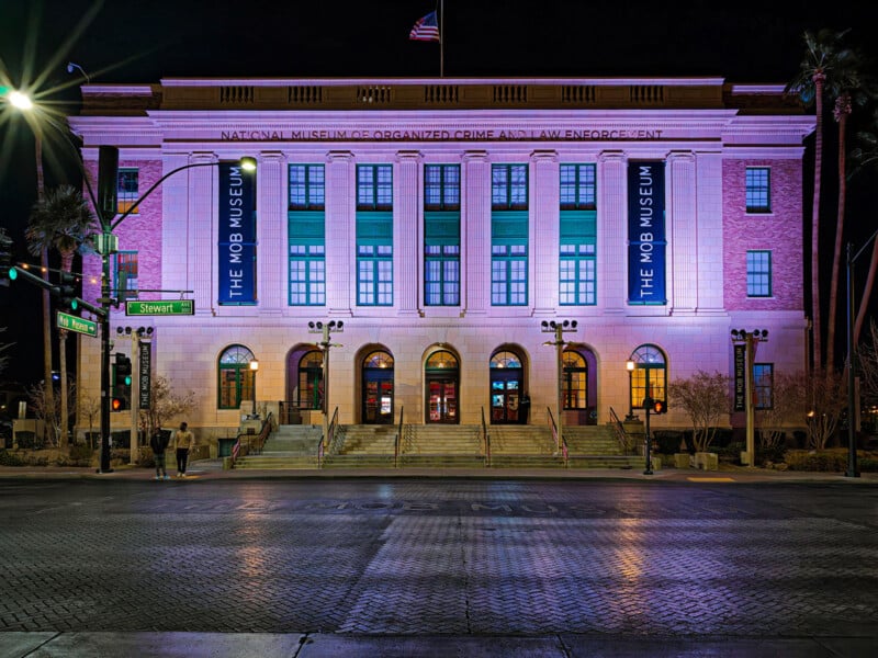 The image shows the exterior of The Mob Museum at night, brightly lit with purple lighting. Large banners on the facade read "The Mob Museum." An American flag flies atop the building.