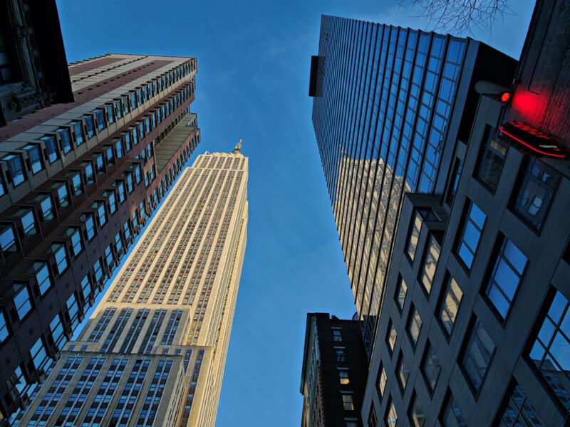 A view looking up at several tall skyscrapers against a clear blue sky. The buildings vary in design, including one with a classic, detailed facade and others with modern glass exteriors. The perspective creates a dramatic, towering effect.