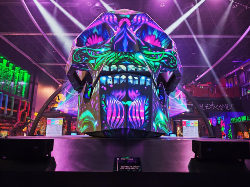 A vibrant, neon-lit skull sculpture with intricate floral and symmetrical designs is on display in a dark room. The colorful lights highlight the patterns, creating a striking visual effect. Surrounding lights add to the immersive atmosphere.