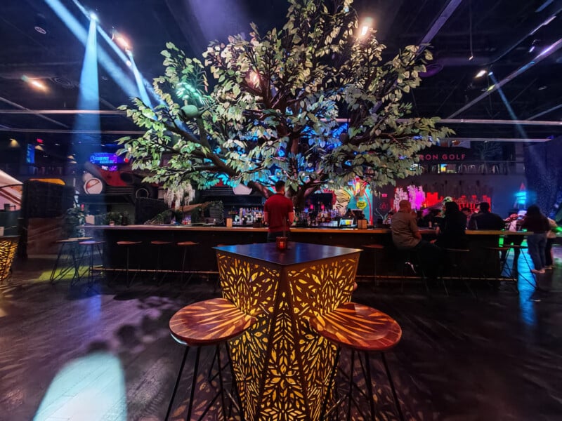 A vibrant night scene in a bar with a large, illuminated artificial tree at the center. People sit at the bar on stools. Decorative lighting casts intricate patterns on the tables. The atmosphere is lively and colorful.