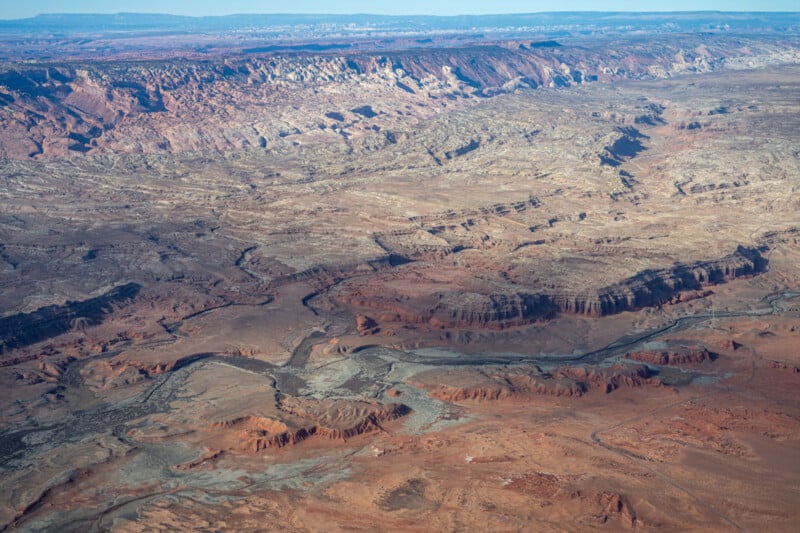 Aerial view of a vast, rugged desert landscape featuring layered rock formations, winding canyons, and sparse vegetation. The terrain is marked by red and brown hues, extending to distant, flat-topped mesas under a clear blue sky.