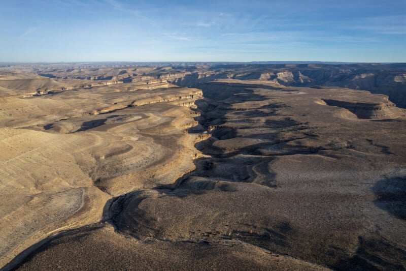 Aerial view of a vast, rugged desert landscape with deep canyons and winding formations under a clear blue sky. The terrain is mostly barren, showcasing layers of sedimentary rock and subtle variations in color and texture.