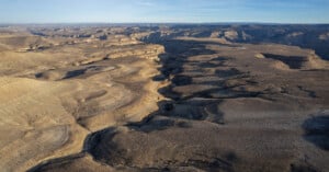 Aerial view of a vast, arid landscape with rugged, layered rock formations and deep canyons under a clear blue sky. The terrain is mostly brown and sparse, showcasing natural erosion patterns.