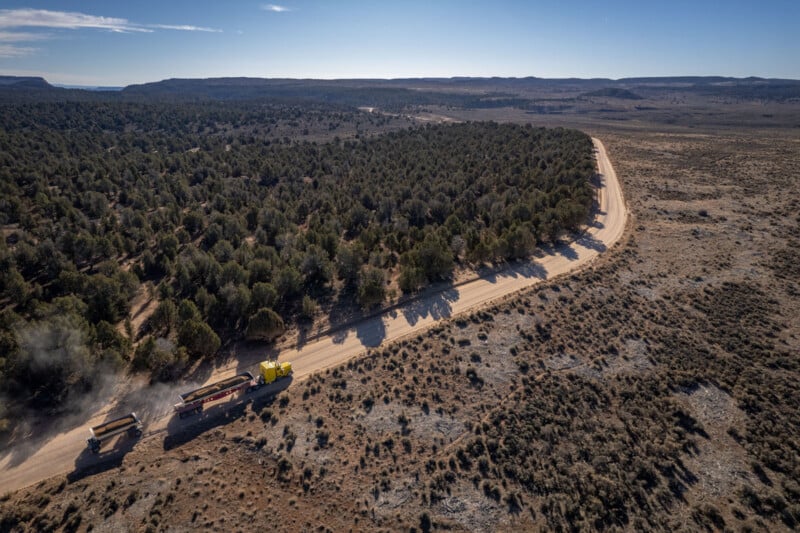 Aerial view of a remote, dusty road winding through a vast, arid landscape. Sparse vegetation and bushy trees dot the terrain. A yellow truck travels along the road, leaving a trail of dust behind. The horizon shows distant hills under a clear sky.