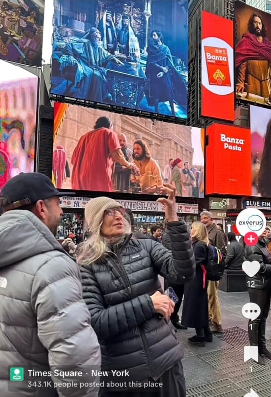 A woman in a gray coat and hat takes a selfie with a man in a black jacket at Times Square, New York. Colorful digital billboards and a bustling crowd fill the background.