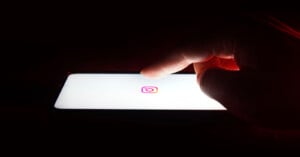 A hand reaching towards a smartphone screen displaying the Instagram logo on a white background, set against a dark backdrop.