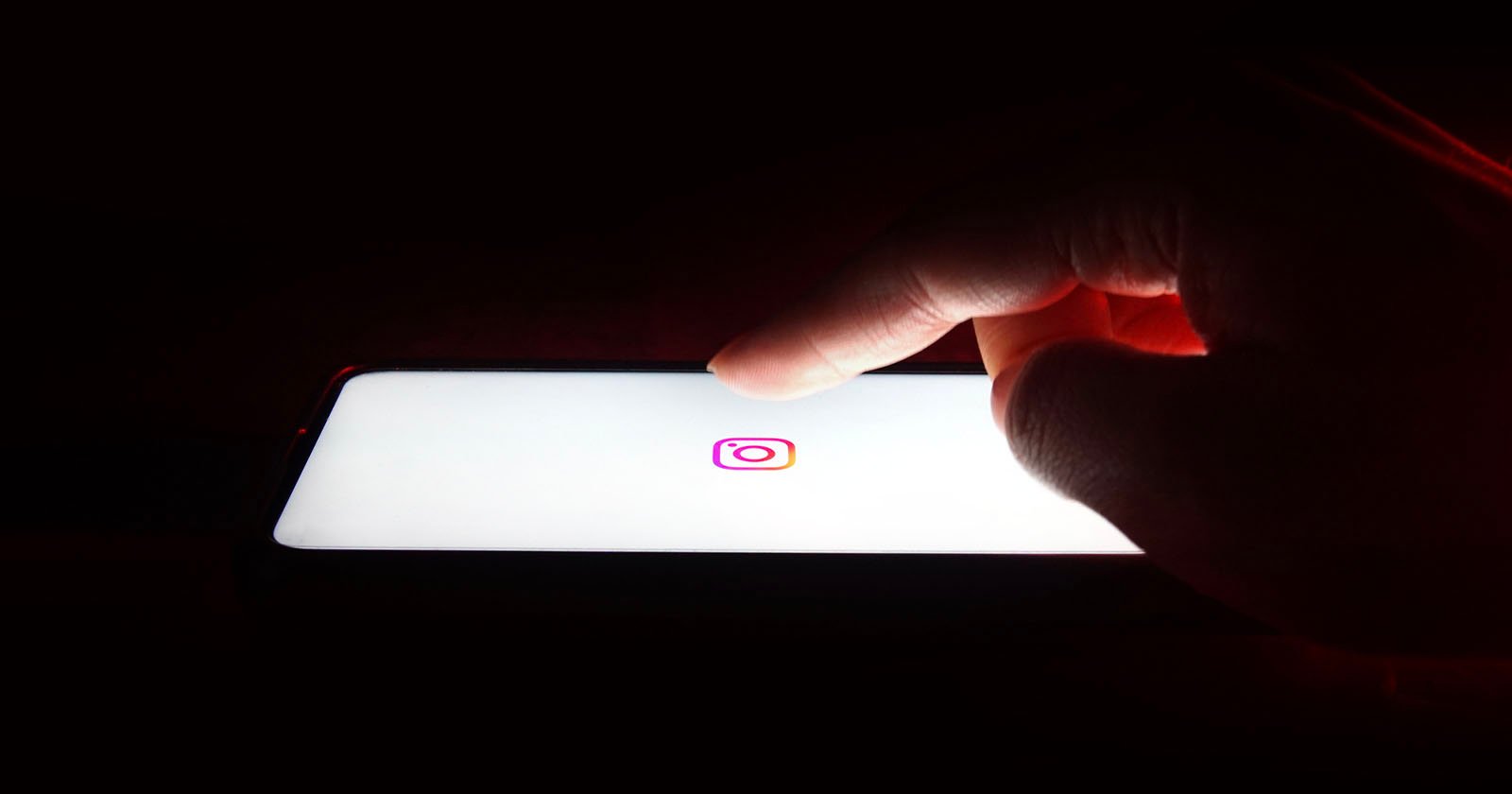 A hand reaching towards a smartphone screen displaying the Instagram logo on a white background, set against a dark backdrop.