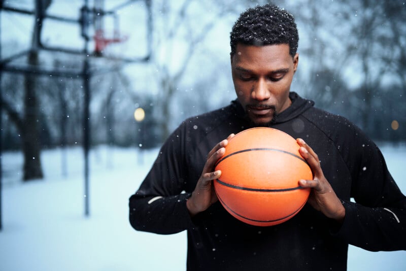 A man in a black hoodie stands outdoors in a snowy setting, holding a basketball. He is looking down at the ball, with a basketball hoop and bare trees visible in the blurred background. Snowflakes are gently falling around him.