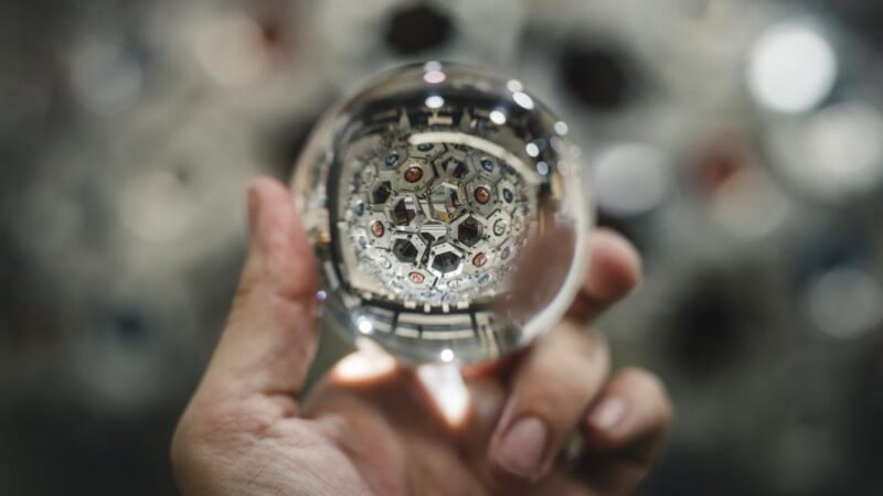 A hand holds a clear glass sphere, reflecting a kaleidoscope of geometric shapes and abstract patterns. The background is blurred, highlighting the sphere's intricate reflections.