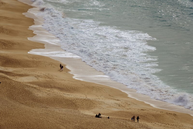 Aerial view of a sandy beach with gentle waves rolling in. A few people stroll along the shoreline and sit on the sand, enjoying the serene atmosphere. The ocean is a light shade of blue under a clear sky.