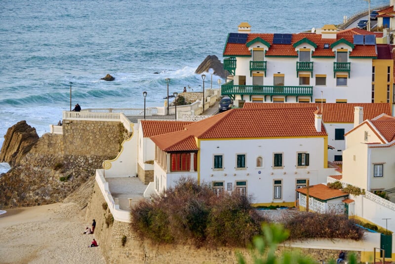 Coastal scene with white and red-roofed buildings overlooking the ocean. Waves crash near a stone wall and a sandy beach. Two people sit on the sand, enjoying the view. Lush greenery in the foreground complements the serene setting.