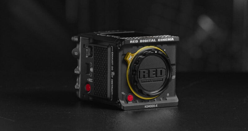 A RED Digital Cinema camera is displayed on a dark surface. The compact, black camera body has multiple controls and a lens mount attached. The brand name "RED" is prominently visible on the front.