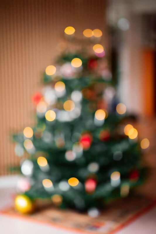 A blurred image of a Christmas tree with soft bokeh lights creating a warm, festive ambiance. The tree's green branches are decorated with various ornaments, including red and gold baubles.