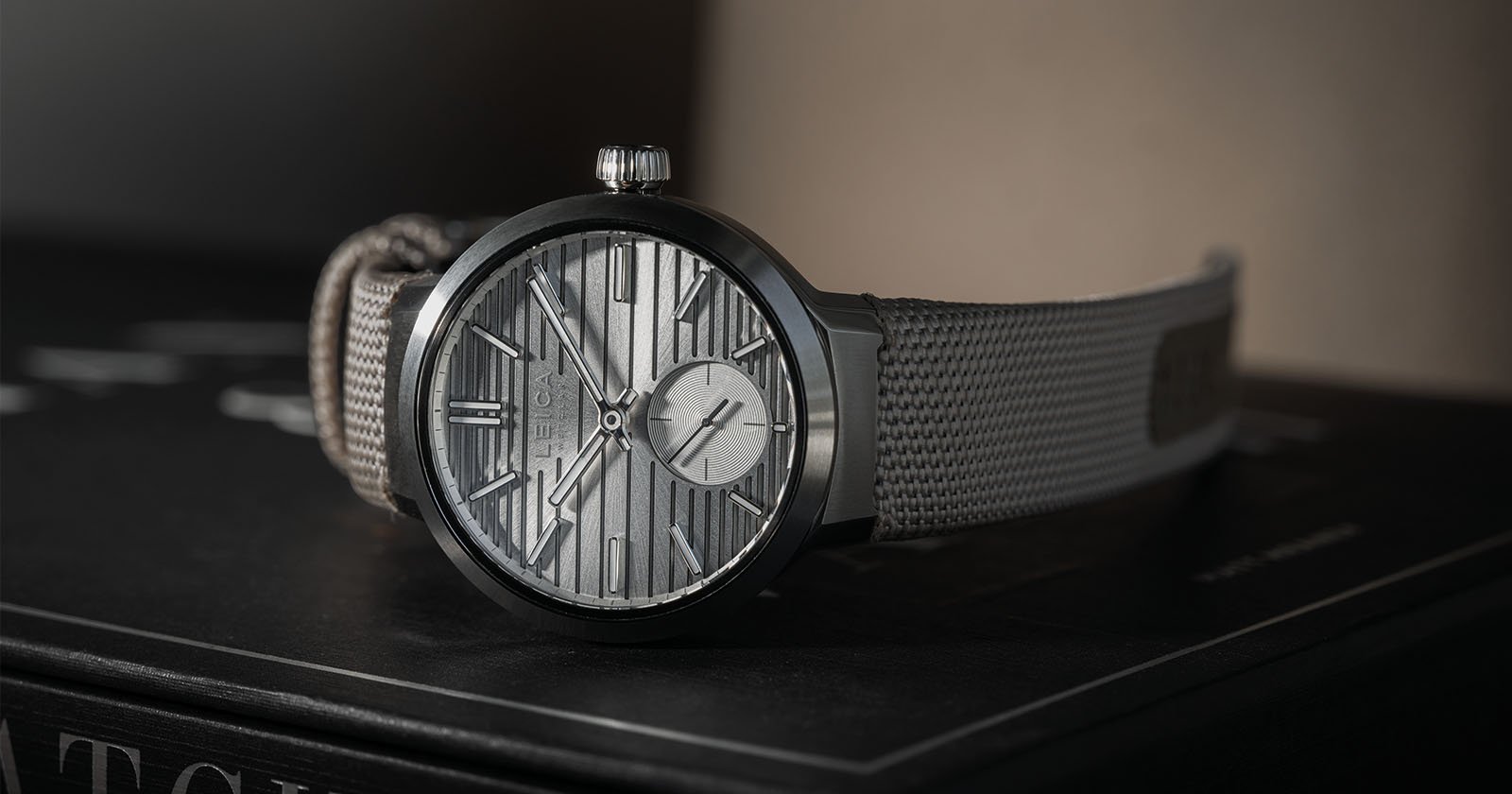 A sleek wristwatch with a textured silver dial featuring geometric patterns and minimalistic hour markers. It has a gray fabric strap and rests on a dark surface with soft lighting highlighting its elegance.