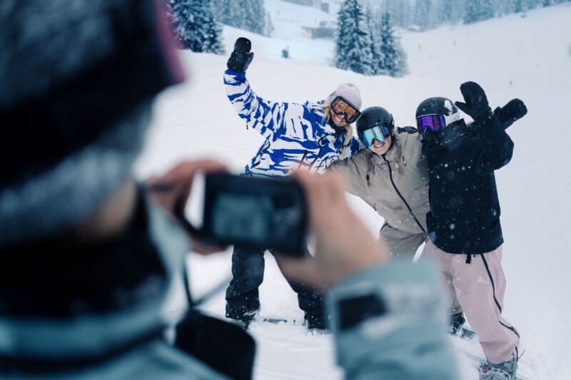 Three people in winter clothing pose together on a snowy slope, smiling and waving, while another person takes their photo. Snow-covered trees are visible in the background.