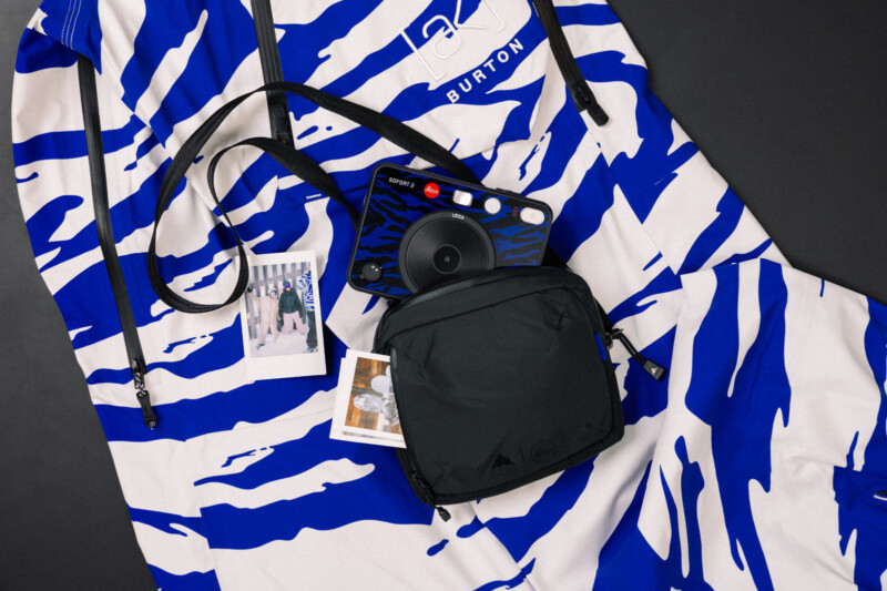 A blue and white patterned hoodie with the logo "Burton" lies on a flat surface. On top are an instant camera with a strap, a small black bag, and two instant photos partially visible.
