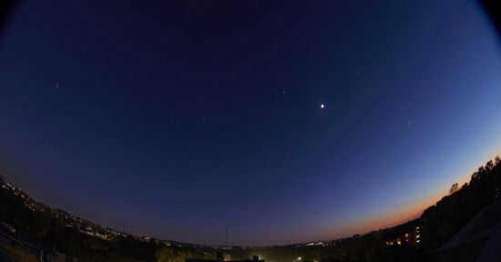 A twilight sky with a bright crescent moon and several stars visible. The horizon glows with the fading light of the setting sun. Below, a landscape of distant city lights and darkened silhouettes of trees is visible.