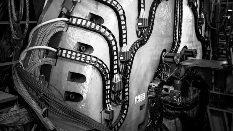 Black and white image of a complex machine with large coils, cables, and chains. The machinery has a metallic surface with a textured pattern and various interconnected components and wires. A warning label is partially visible.