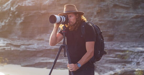 A person with long hair, wearing a hat and a black backpack, is using a camera on a tripod. They are outdoors, standing in front of a rocky landscape. The lighting is warm and the setting suggests a natural environment.