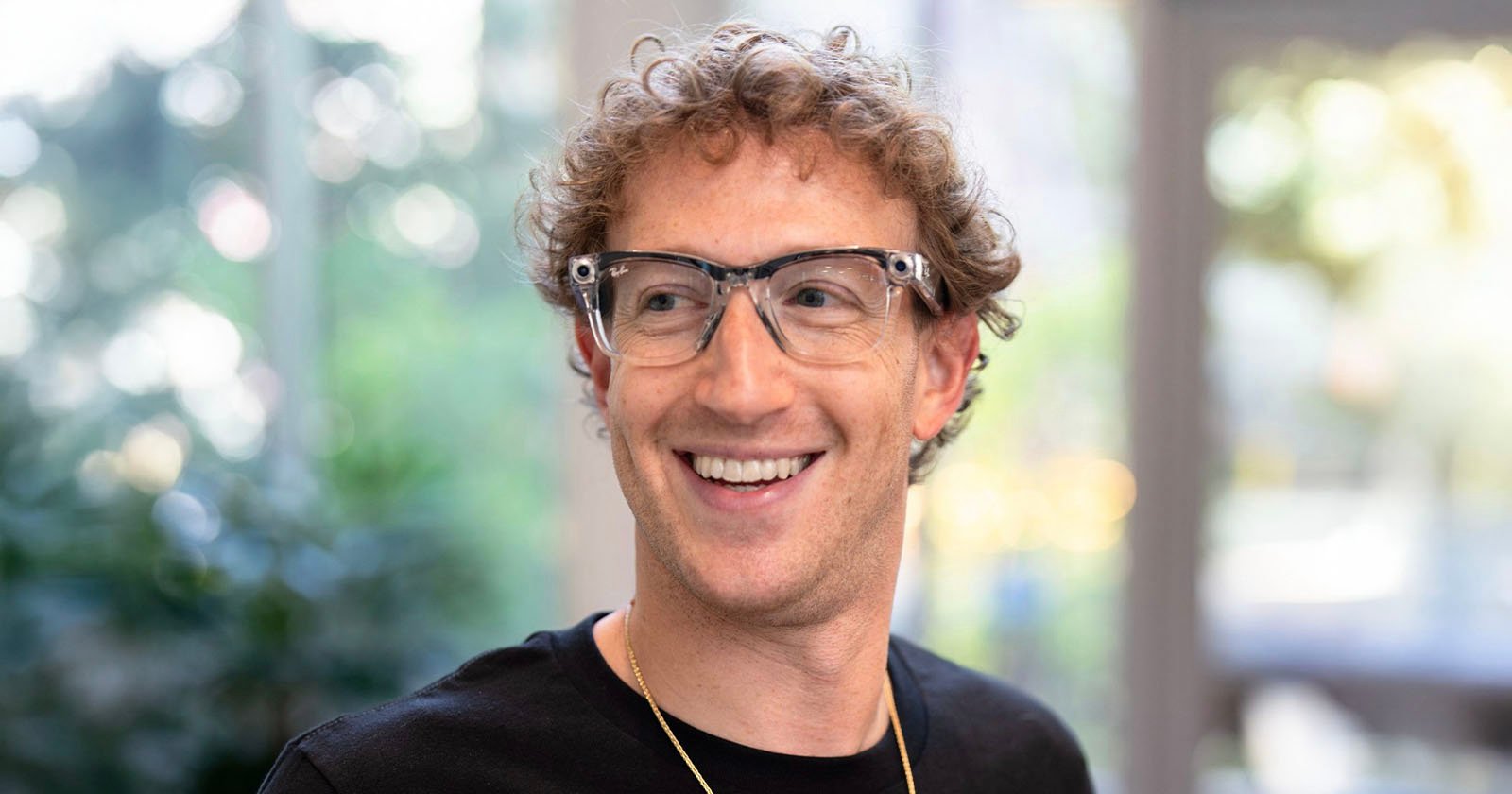 A person with curly hair is smiling while wearing transparent glasses and a black shirt. They are standing indoors, with a blurred background featuring greenery and large windows.