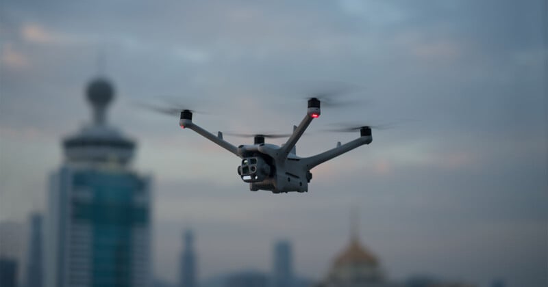 A drone with six propellers flies in the sky against an urban cityscape background featuring modern buildings and a hazy cloud cover.