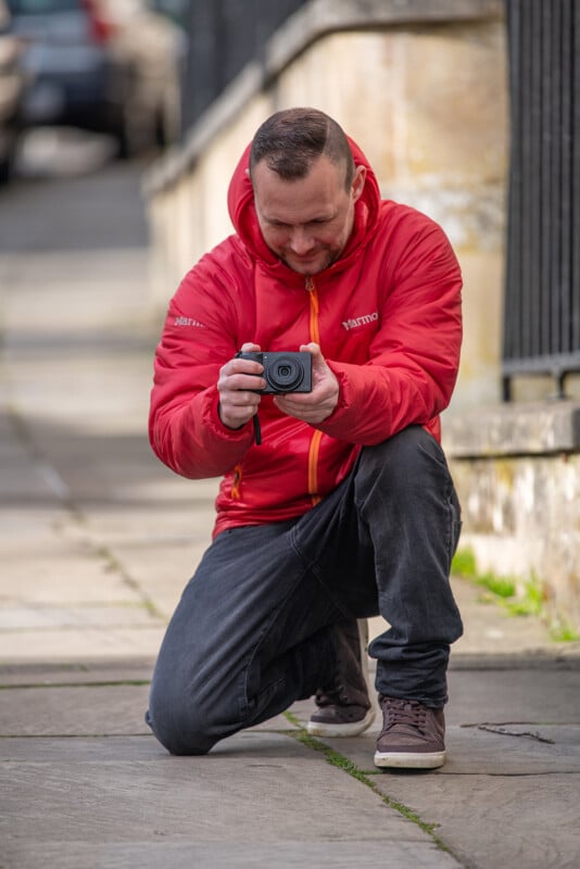 A person in a red jacket crouches on a sidewalk, holding a camera and focusing on a shot. The background is slightly blurred, showing a stone building and part of a street.