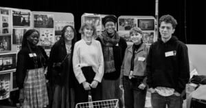 A group of six people standing together, smiling at the camera in a room with display boards filled with photos in the background. Some wear name tags and casual clothing, while others sport scarves and glasses. Black and white image.